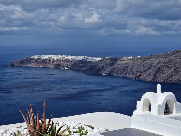 A wide, storm-lit view along the caldera cliffs toward a distant whitewashed village under heavy clouds