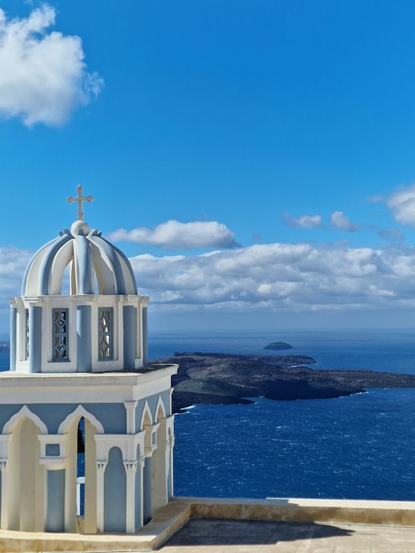 A whitewashed church bell tower in Imerovigli, with the dark bulk of Skaros Rock rising from the caldera behind
