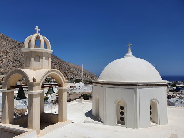 A whitewashed church dome and bell tower above a hillside village, with the sea visible in the distance