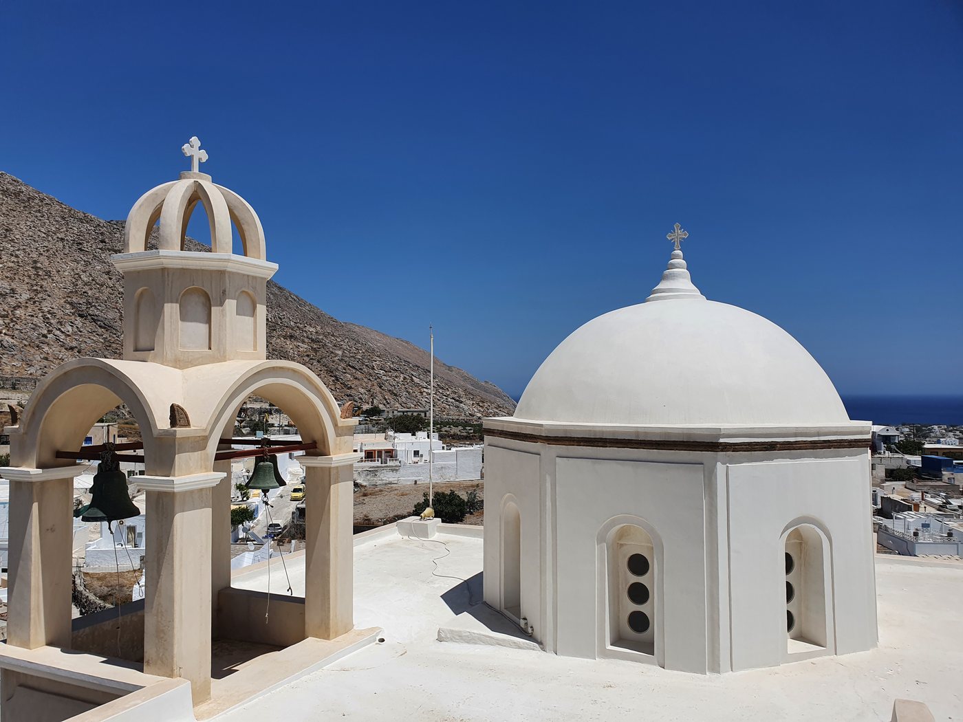 A whitewashed church dome and bell tower above a hillside village, with the sea visible in the distance