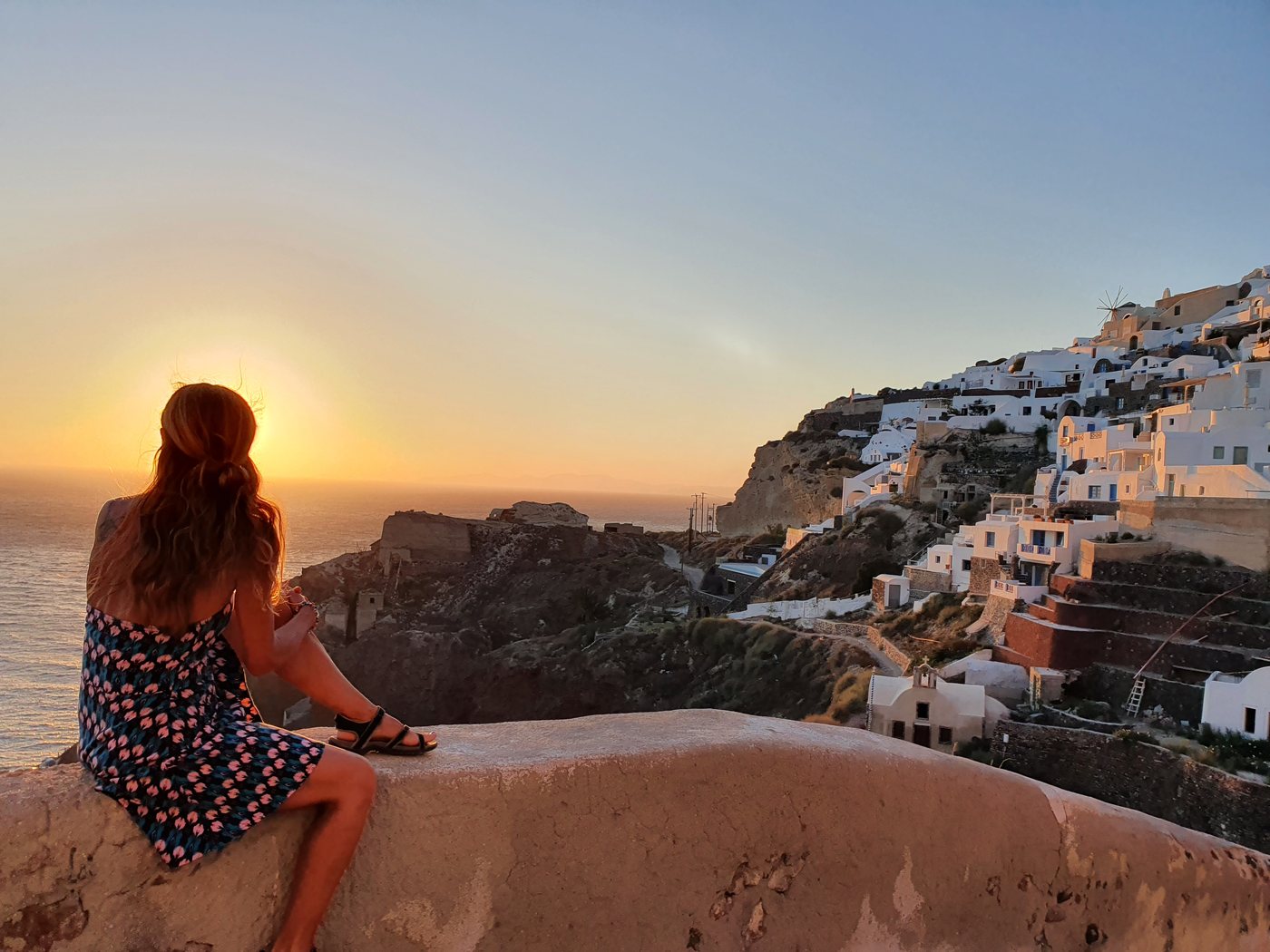 A visitor watches the sun go down from Oia's castle viewpoint, with the village and a windmill spread along the cliff