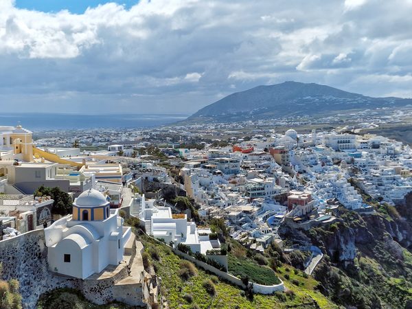 Fira town cascading down the caldera cliff, seen from above on a cloudy day with the island's highest peak behind it