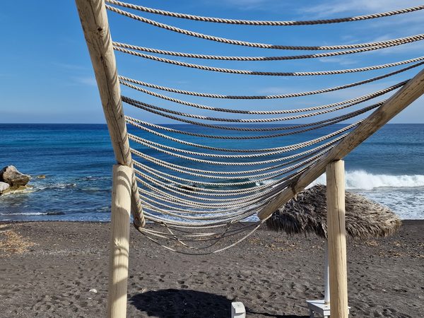 A wooden hammock frame and straw beach umbrella on the black sand at Perivolos, with waves breaking behind
