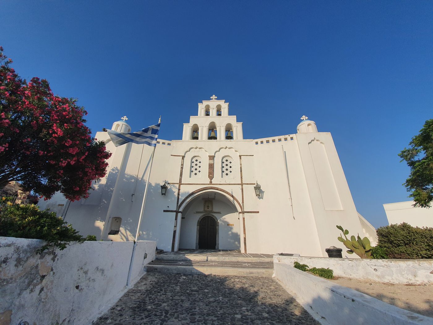 A fortress-like whitewashed church with a triple-bell arch in Pyrgos, a Greek flag flying beside its stone entrance path