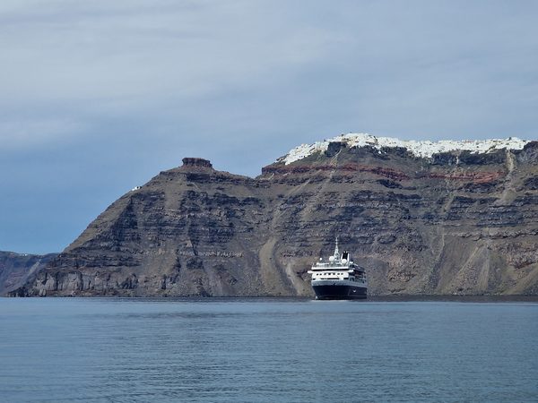 A small cruise ship on the caldera waters below Santorini's striped volcanic cliffs, with the whitewashed clifftop town above