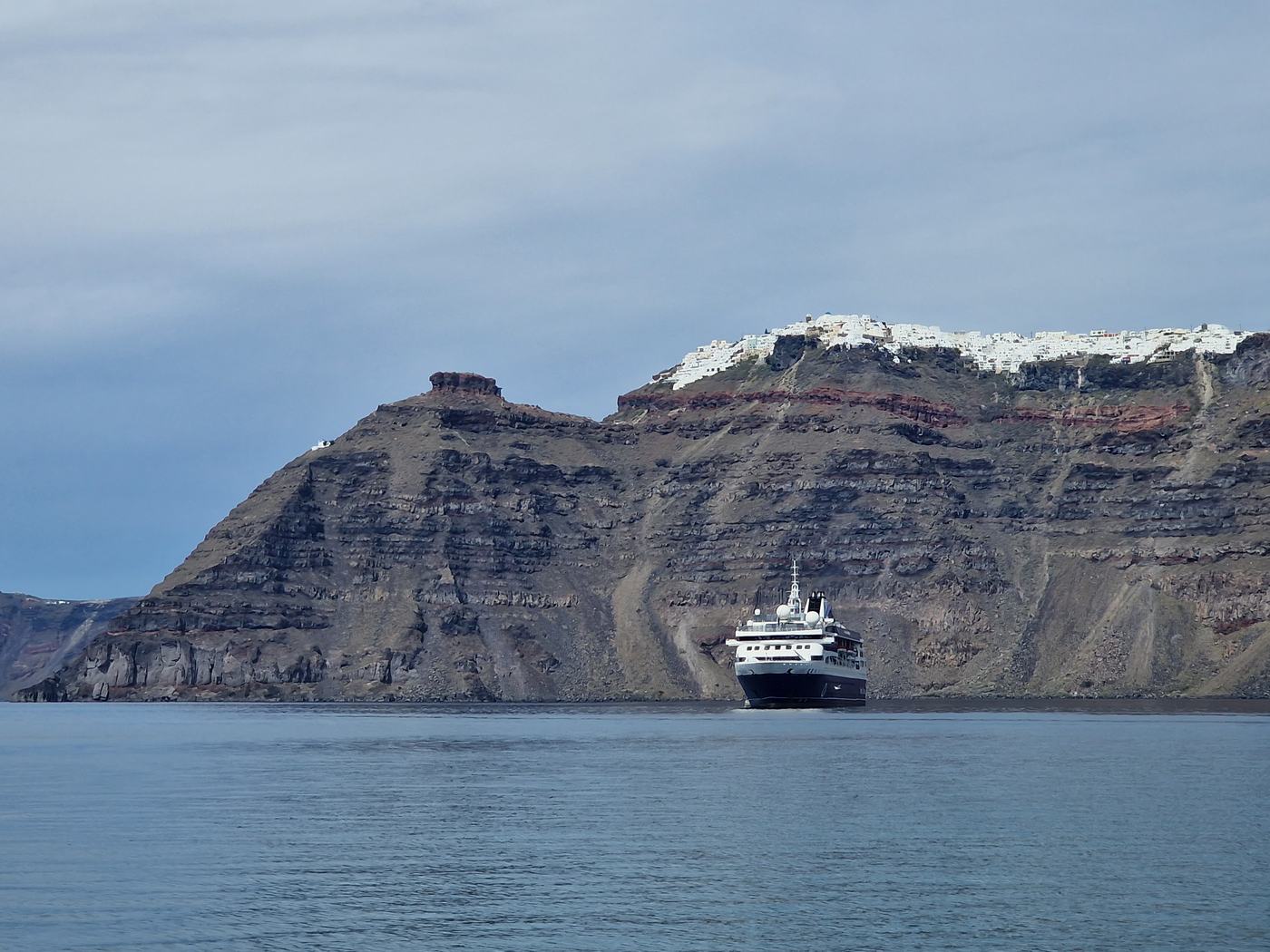 A small cruise ship on the caldera waters below Santorini's striped volcanic cliffs, with the whitewashed clifftop town above