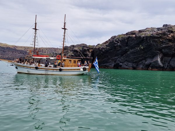 A wooden tour boat anchored in the sheltered cove at Palea Kameni, its mineral-stained water a distinct green against the black volcanic cliffs