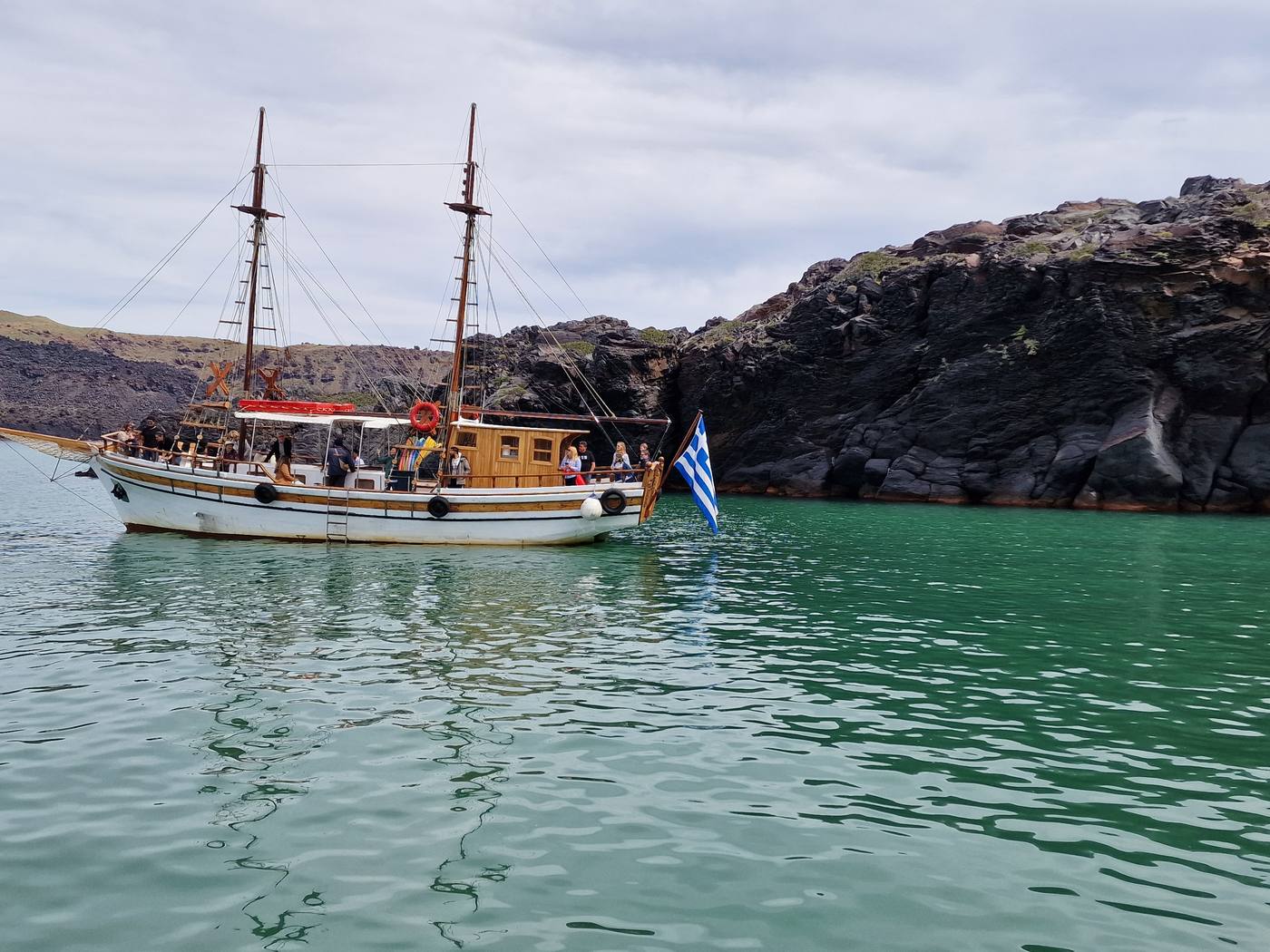 A wooden tour boat anchored in the sheltered cove at Palea Kameni, its mineral-stained water a distinct green against the black volcanic cliffs