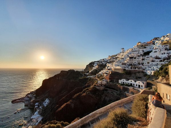The sun setting over the sea beside Oia, its whitewashed houses glowing gold and small boats anchored in the water below the cliffs
