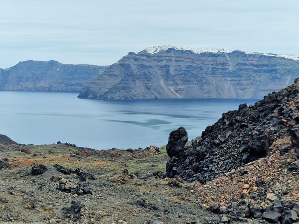 Black volcanic rock on the Nea Kameni crater path with the Santorini caldera and clifftop village in the distance