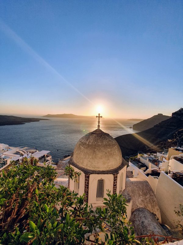 A church dome and cross silhouetted against the sun setting over the Santorini caldera
