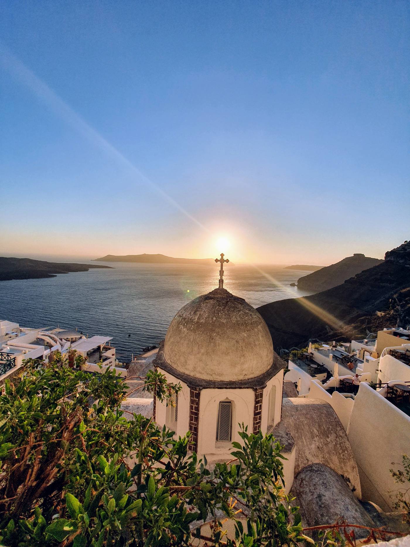 A church dome and cross silhouetted against the sun setting over the Santorini caldera